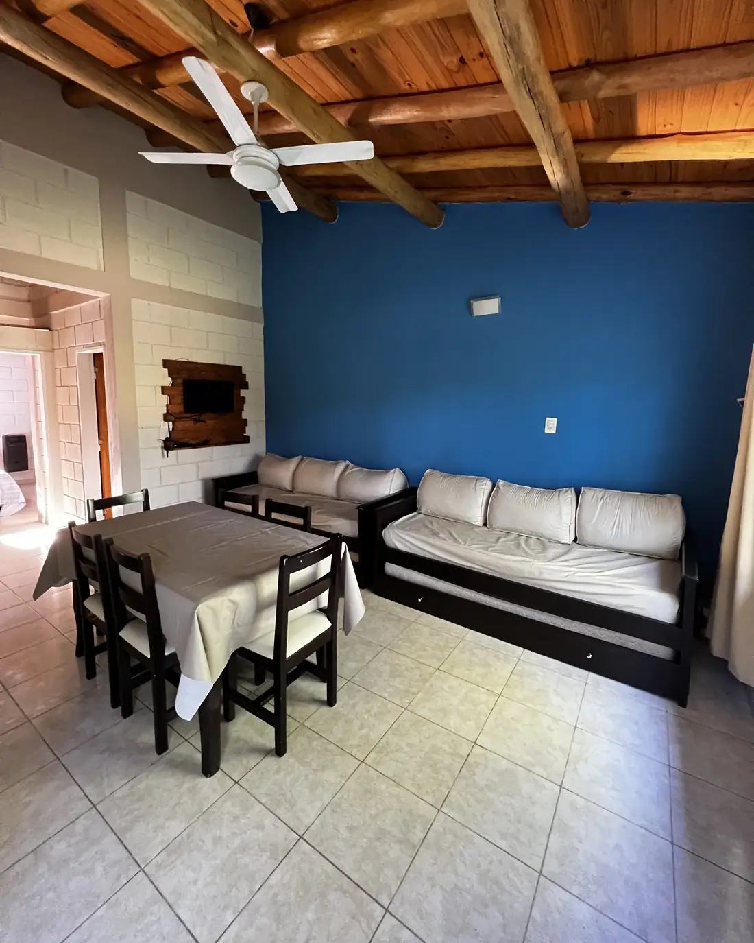 Living comedor de una cabaña con techo de madera, mesa, sofás cama y pared azul
